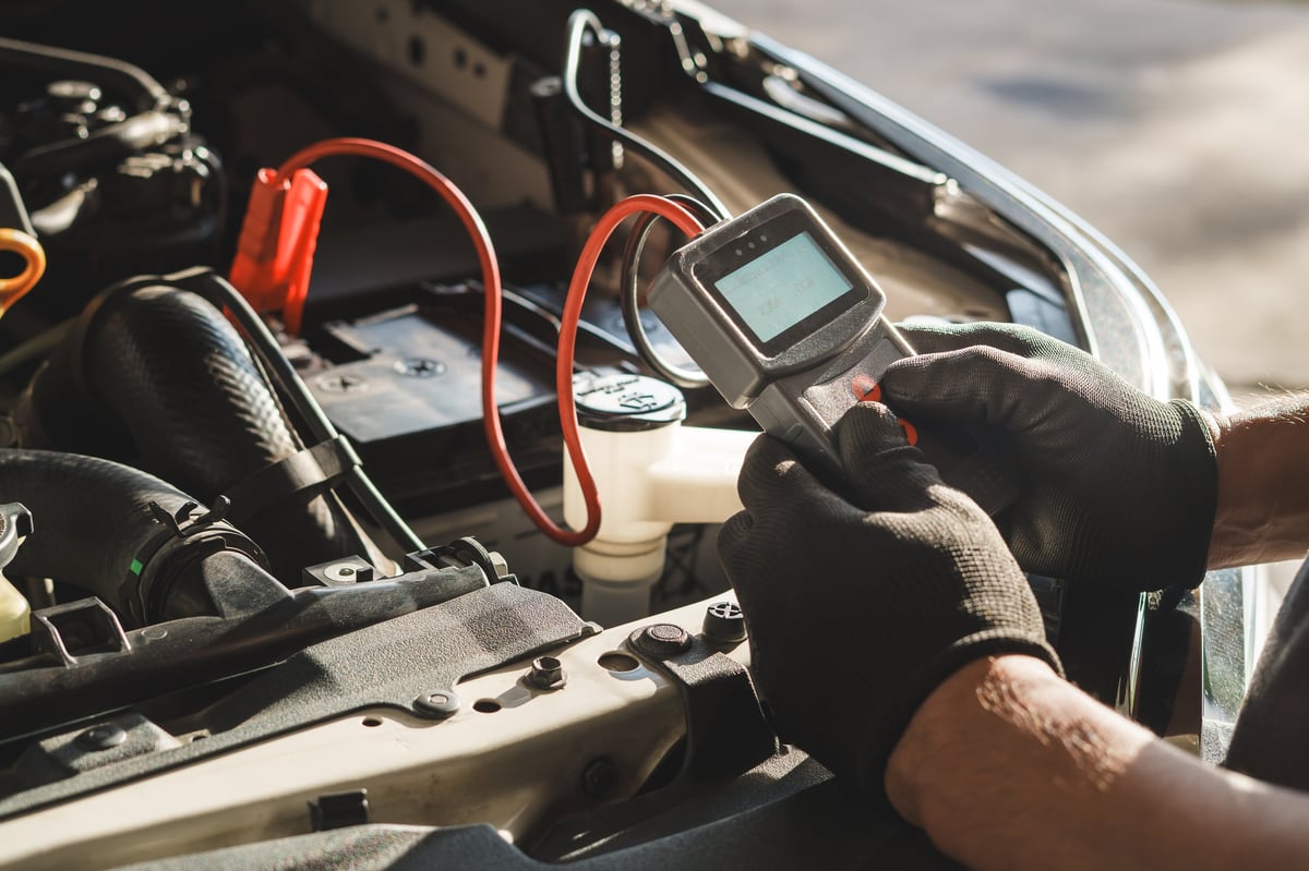 Professional car mechanic using a digital battery tester to inspect vehicle performance during routine maintenance. Automotive technician wearing gloves performing diagnostics under the hood in a repair shop.